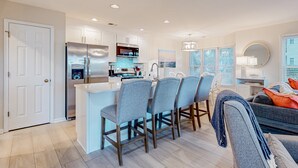 A modern kitchen with a central island, bar stools, and a dining area with a sofa. - Ev's Escape,201A: Modern condo with sunroom - dog and snowbird-friendly (Sunset Beach)
