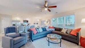 A modern living room with a sofa, armchair, and coffee table. There is a ceiling fan, a dining area with chairs, and a kitchen in the background. - Ev's Escape,201A: Modern condo with sunroom - dog and snowbird-friendly (Sunset Beach)
