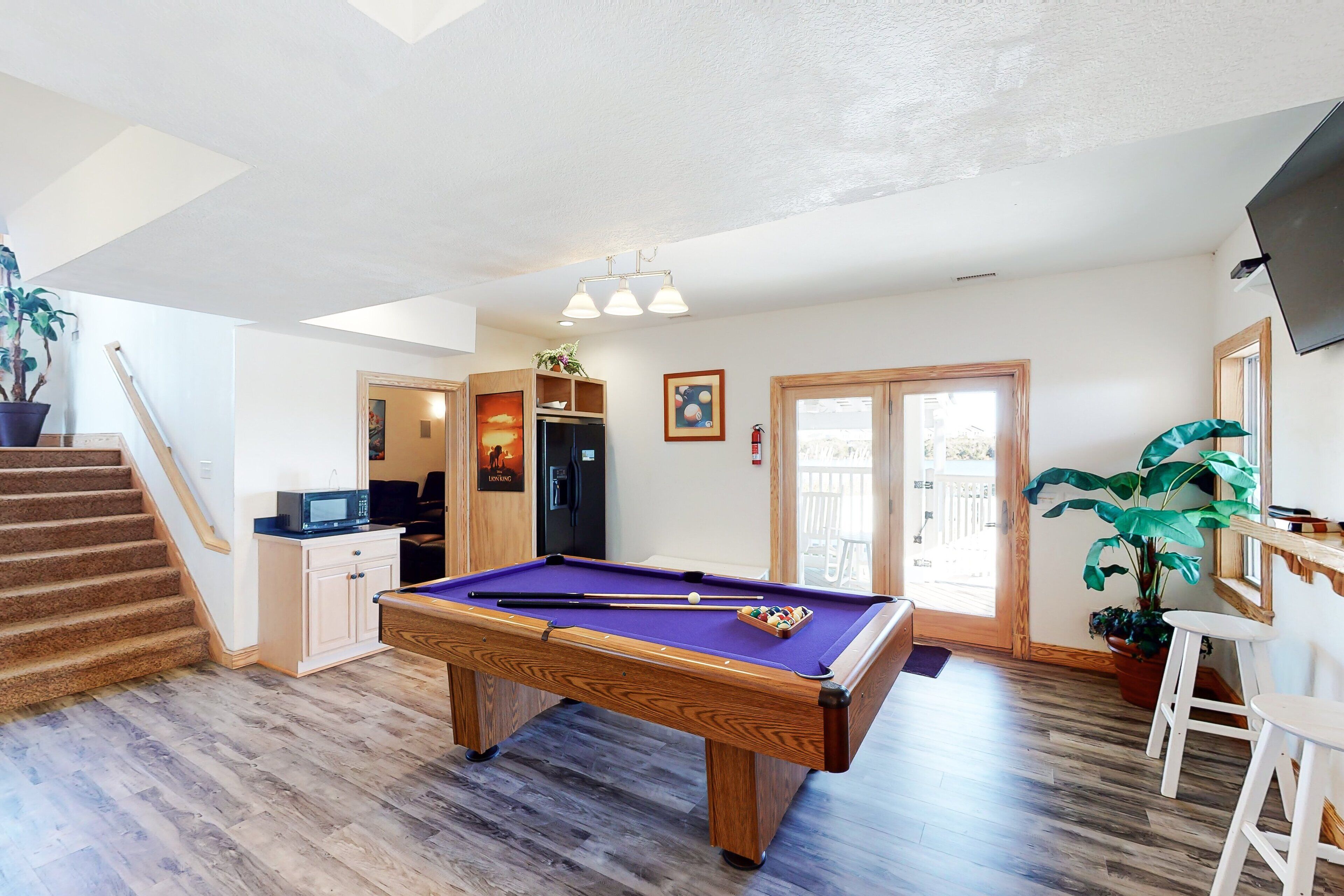 A pool table in a room with wooden flooring, a microwave, a refrigerator, and a television mounted on the wall.