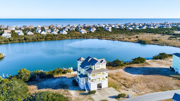 A coastal residential area with a large body of water, a prominent multi-story house with a balcony, and a dense cluster of houses in the background. - Lakefront, dog-friendly home with W/D, private pool & hot tub - close to beach (Avon)