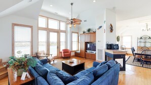 A spacious living room with a blue sofa, a wooden coffee table, and a television. There is a kitchen area with wooden cabinets and a dining table with chairs. - Lakefront, dog-friendly home with W/D, private pool & hot tub - close to beach (Avon)