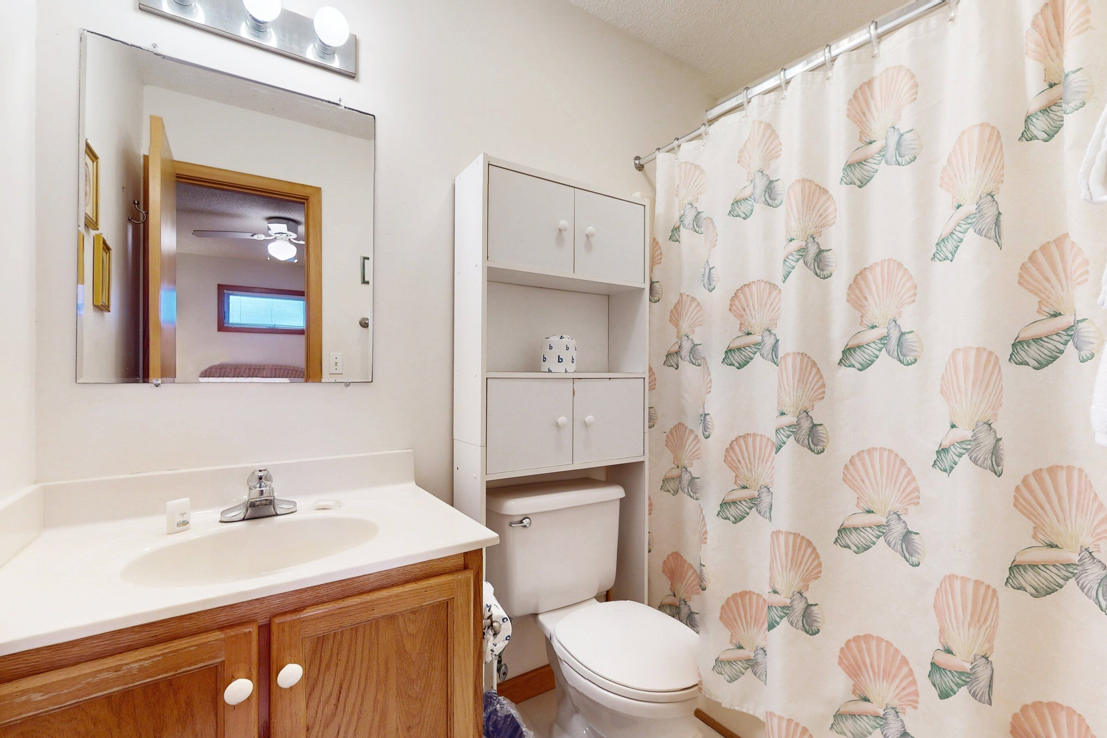 A bathroom with a white sink, wooden cabinets, a toilet, and a shower curtain with a shell pattern.