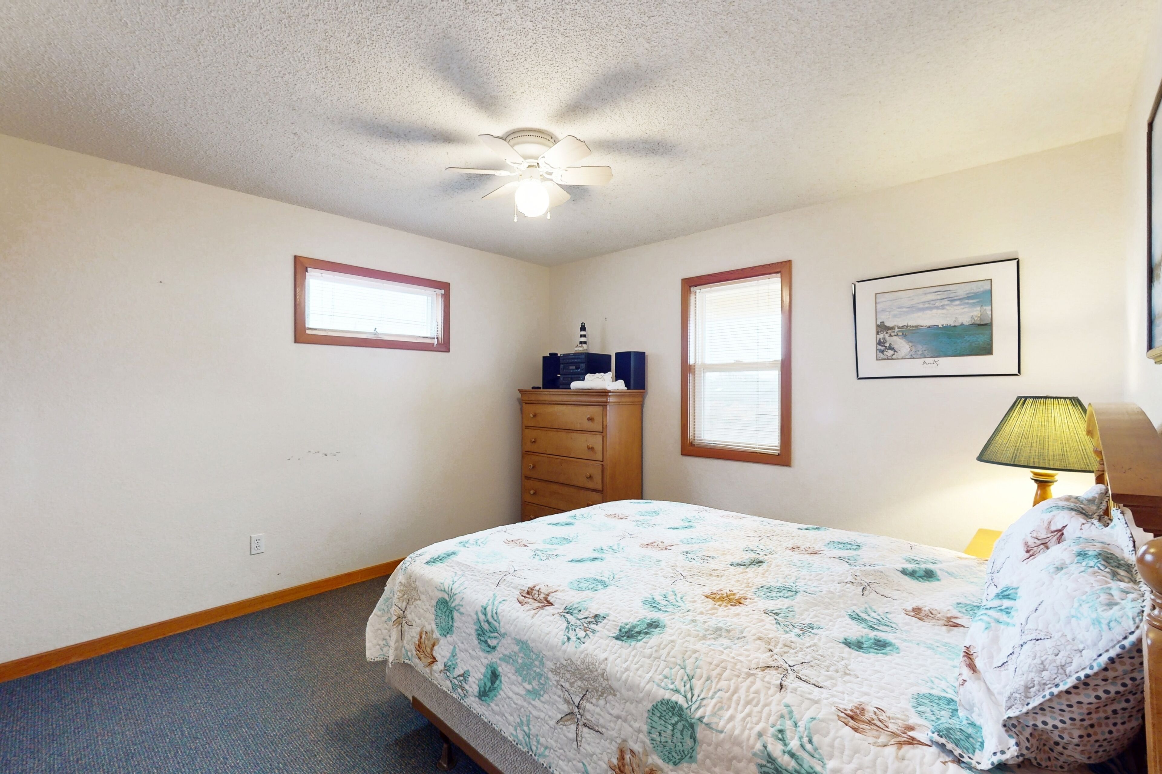 A bedroom with a bed, dresser, ceiling fan, windows, and a lamp.