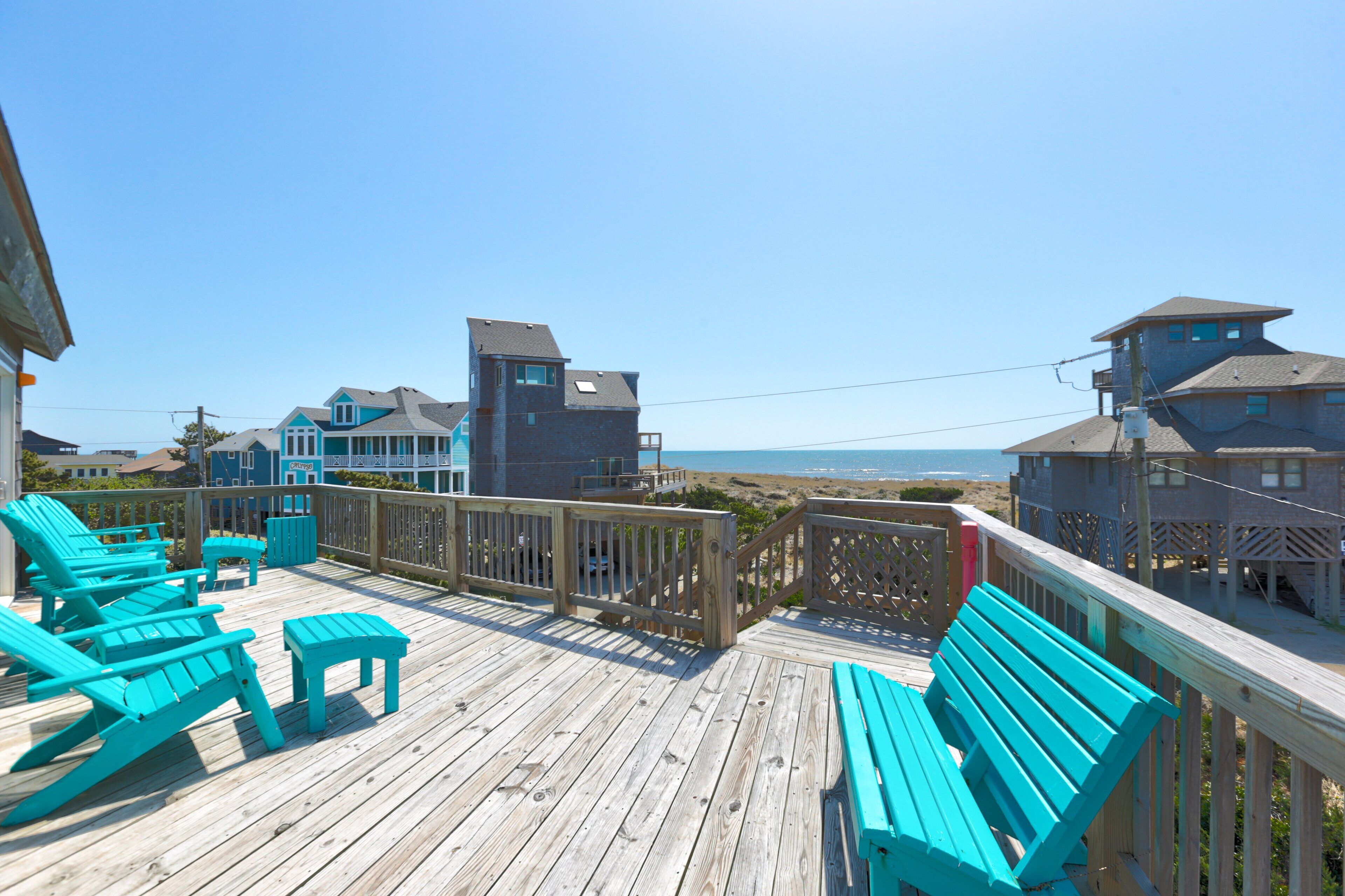 A wooden deck with turquoise chairs and a bench, overlooking a beach and buildings.