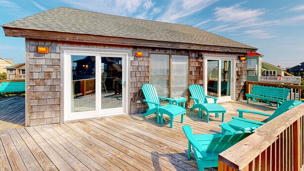 Une terrasse en bois avec des chaises et des tables turquoise, un patio couvert et un bâtiment doté de grandes portes-fenêtres coulissantes.