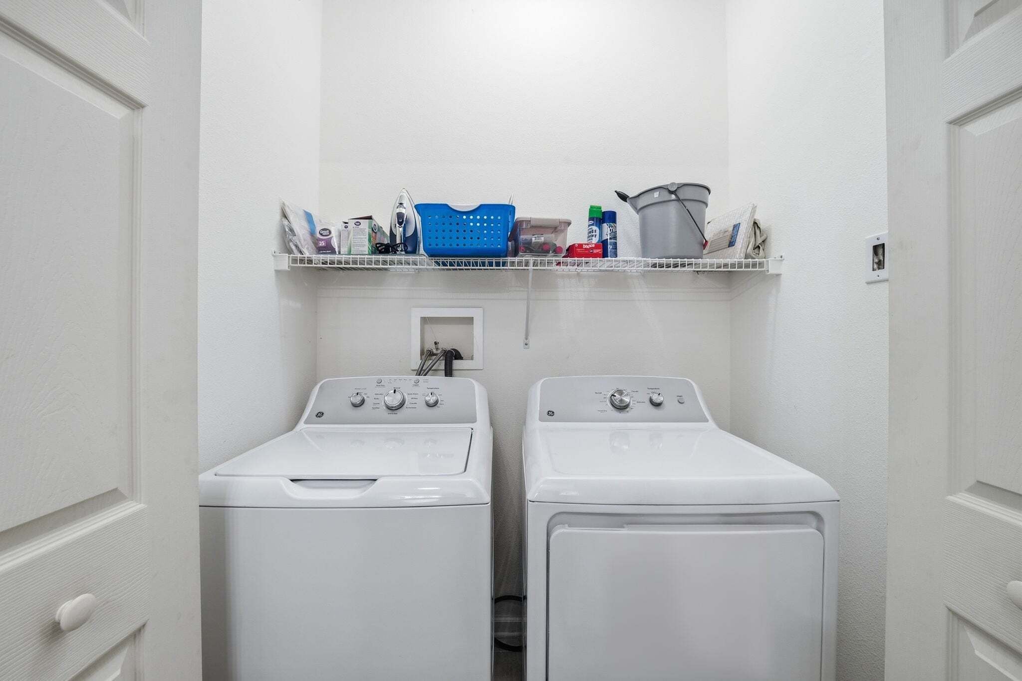 A laundry room with two front-loading washers and dryers, a shelf above with various laundry supplies, and a small window.
