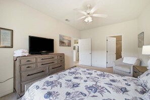 Une chambre à coucher comprenant un lit, une commode, un téléviseur, un ventilateur de plafond et une porte donnant sur une autre pièce.