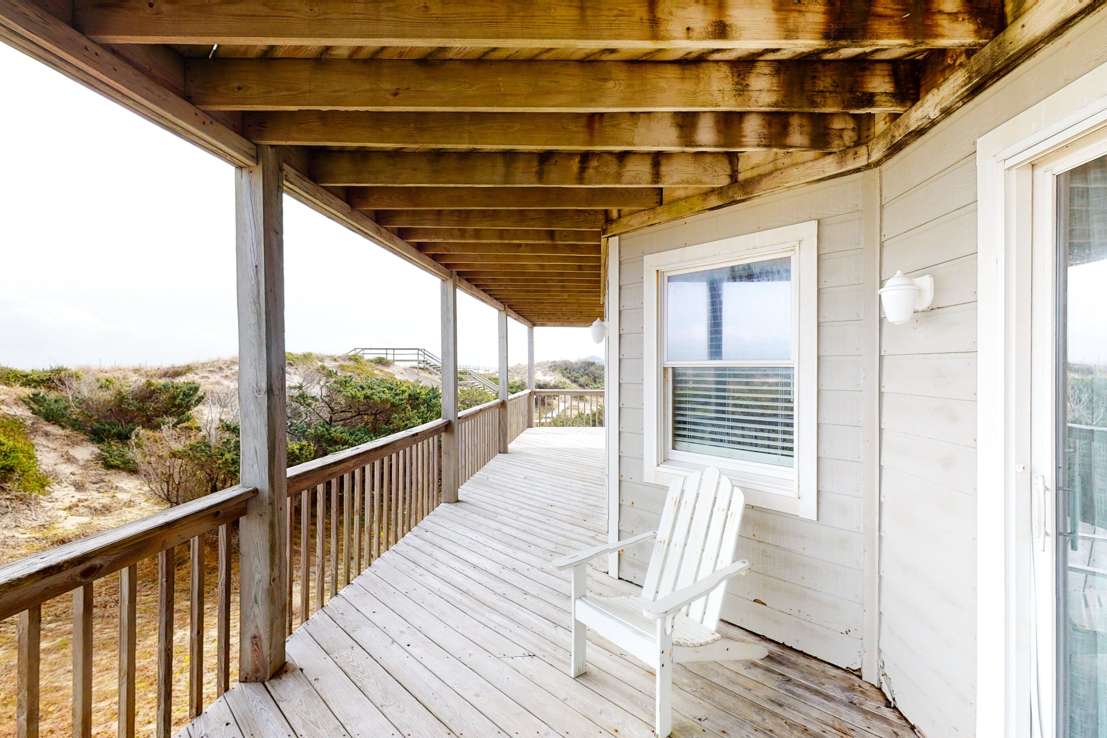 A wooden deck with a white chair and a window overlooking a beach.