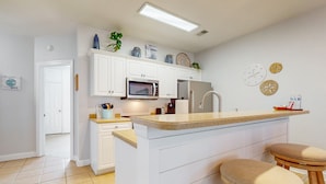 A modern kitchen with white cabinetry, a central island with stools, a microwave, and a stainless steel refrigerator. - SB72A&B: Coastal Condo with golf course view and shared pool access (Sunset Beach)