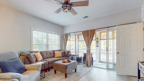 A spacious living room with a grey sofa, a blue armchair, and a wooden coffee table. A ceiling fan and a large window with curtains are visible. - SB72A&B: Coastal Condo with golf course view and shared pool access (Sunset Beach)