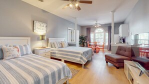 A well-lit bedroom with two beds, a ceiling fan, a dining area with a table and chairs, and a kitchenette. - SB72A&B: Coastal Condo with golf course view and shared pool access (Sunset Beach)