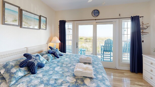 A bedroom with a bed, a ceiling fan, a view of the balcony, and a window with curtains. - Inviting beachfront condo featuring hot tub & pool, central AC, elevator, & deck (Hatteras)