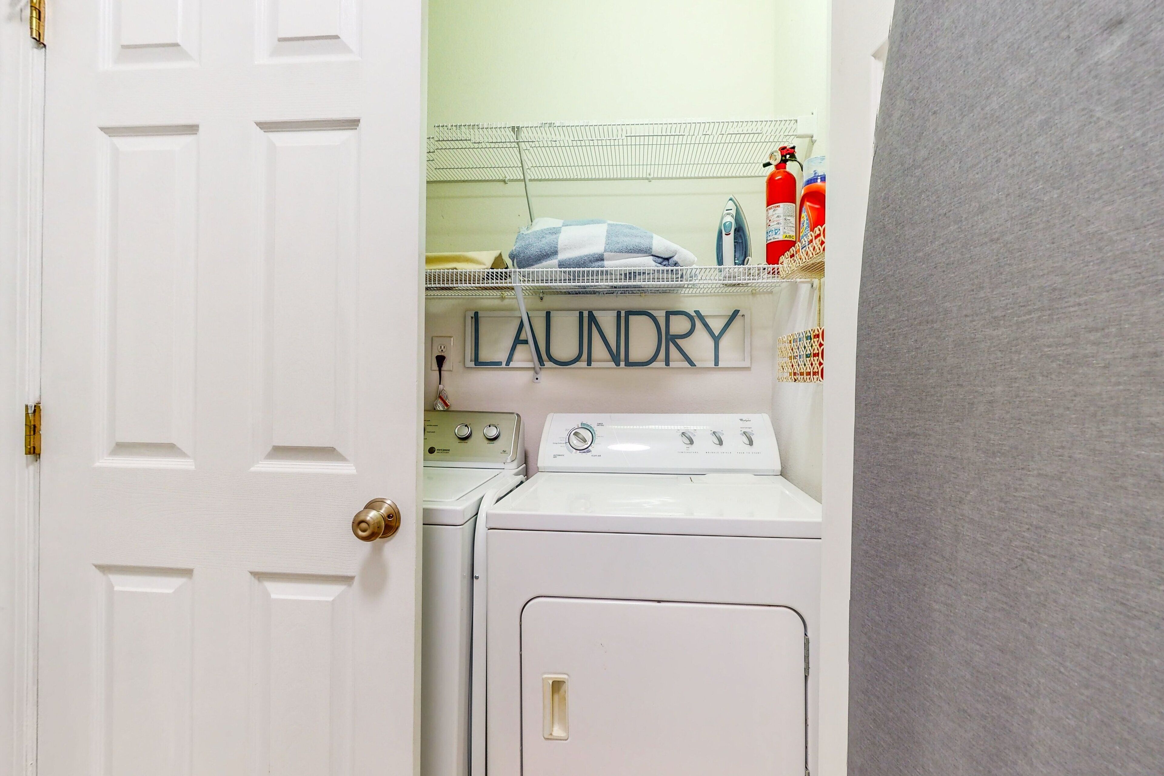 A laundry area with a white door, a washing machine, an iron, and a shelf with laundry supplies.