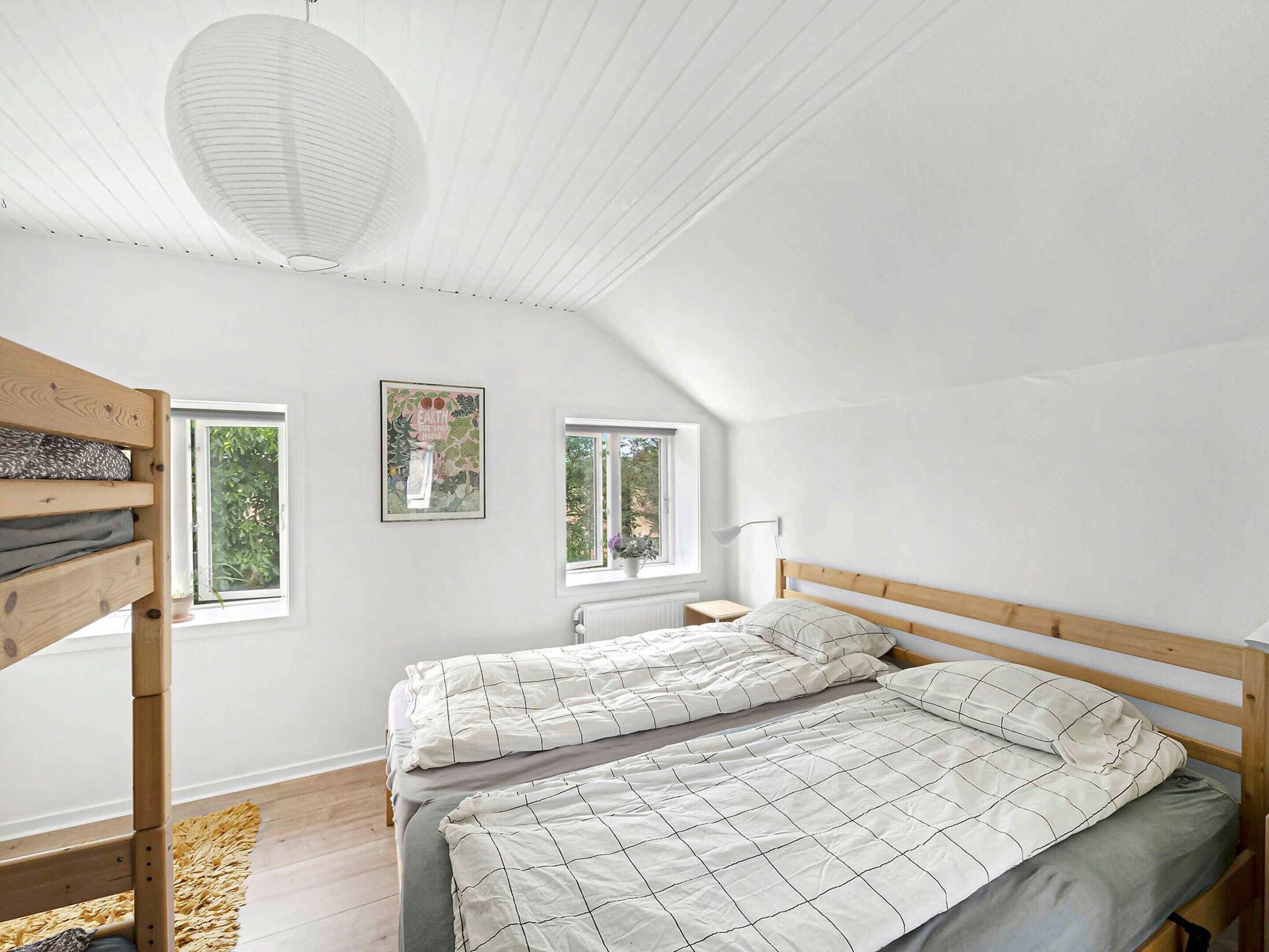 Une chambre avec un plafond en pente, deux lits avec du linge de lit blanc, un lit superposé en bois, deux fenêtres donnant sur la verdure et un ventilateur de plafond blanc.