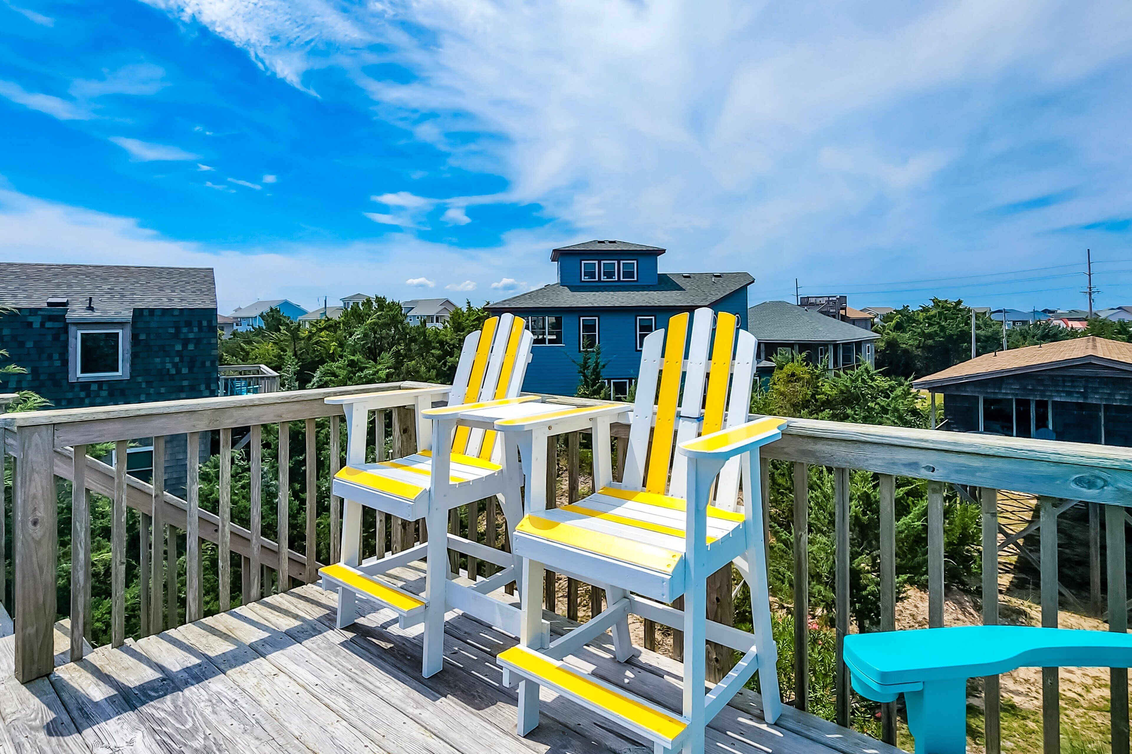 Une terrasse en bois avec des fauteuils Adirondack blancs et jaunes et un banc turquoise.