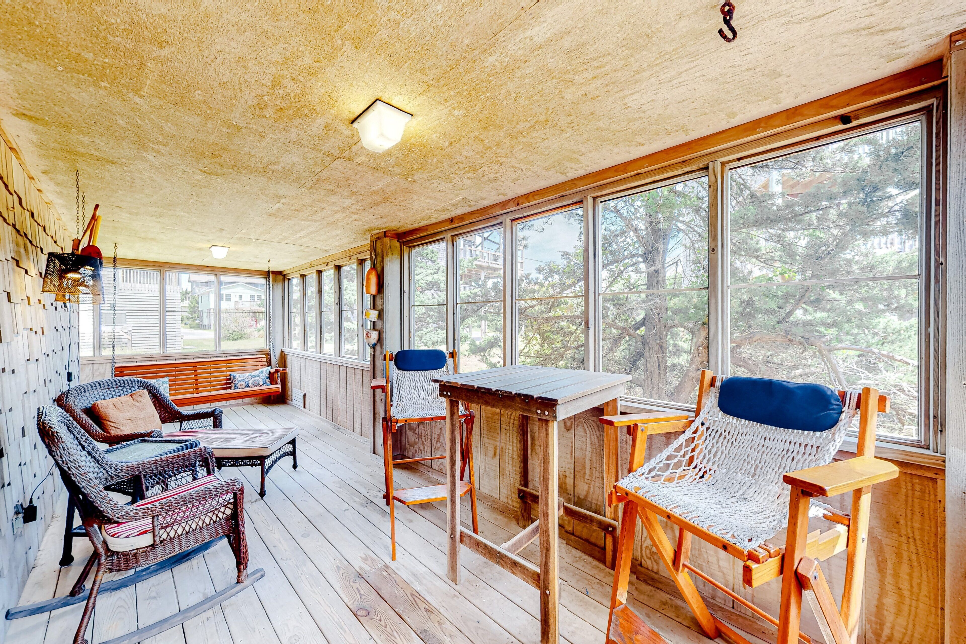 A covered porch with wicker furniture, a wooden table, and large windows offering a view of trees.