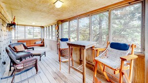 Varanda coberta com móveis de vime, uma mesa de madeira e grandes janelas com vista para árvores.