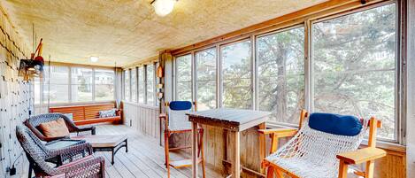 A covered porch with wicker furniture, a wooden table, and large windows offering a view of trees.