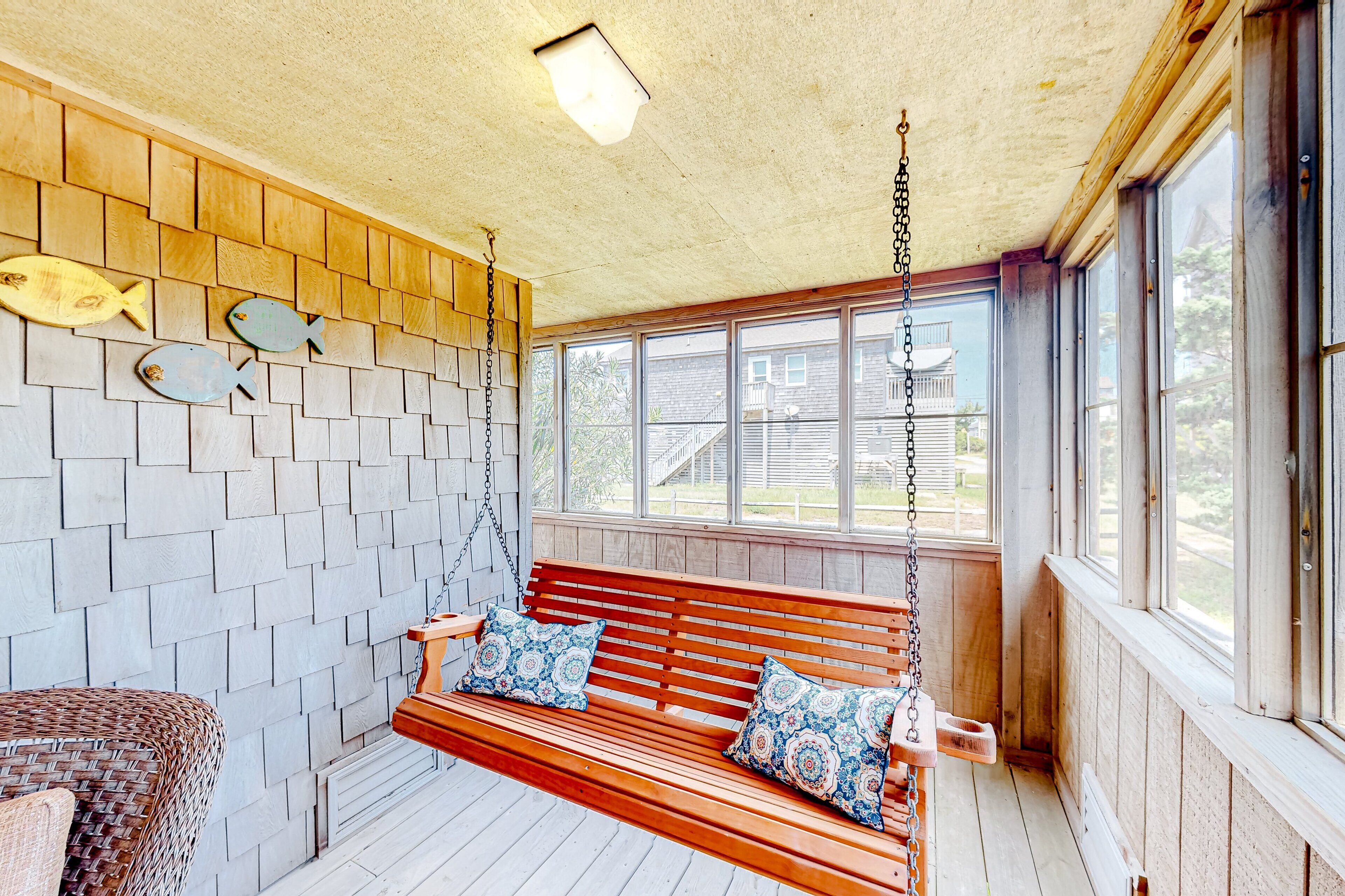 A wooden bench with patterned cushions in a covered porch area with a view of a house through large windows.