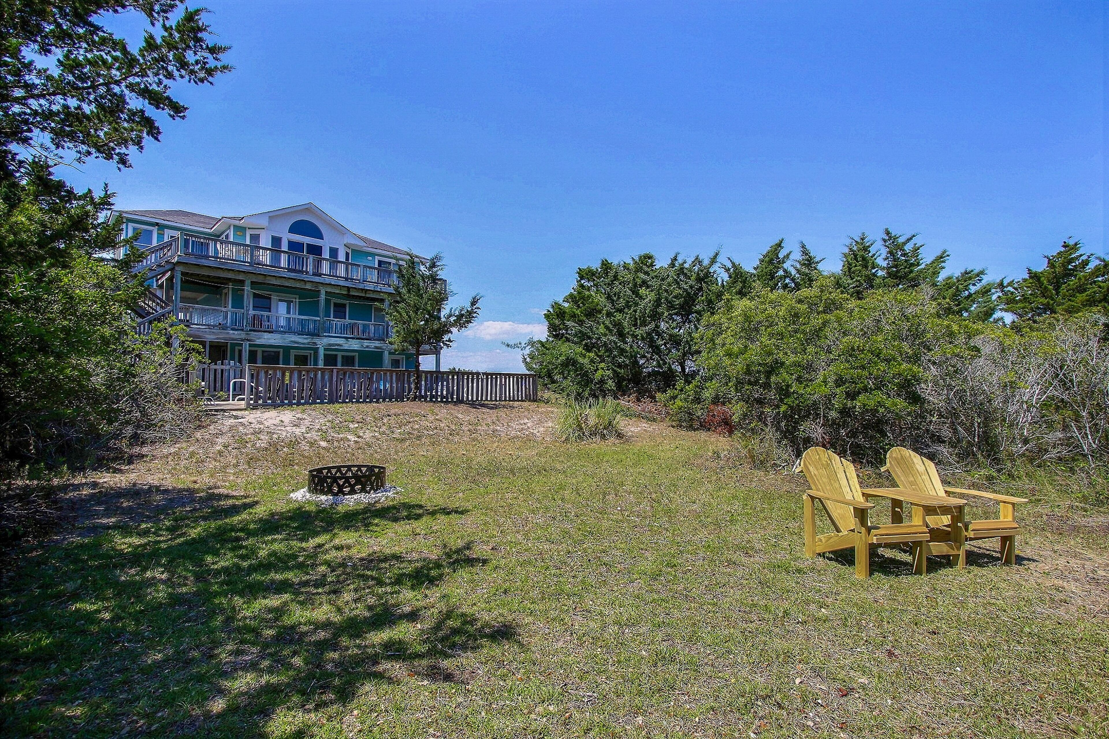 A two-story house with a balcony, surrounded by a wooden fence and trees. There are two Adirondack chairs in the foreground.