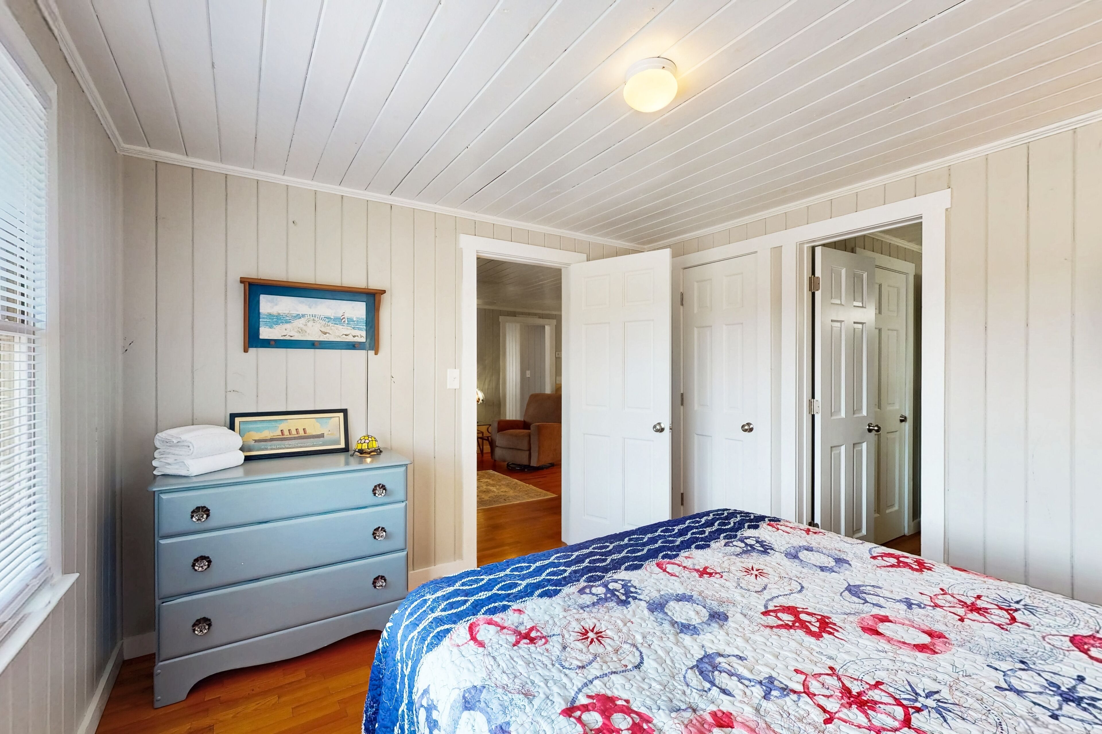 Ein Schlafzimmer mit einem Bett, einer Kommode, einem gerahmten Bild und einer offenen Tür zu einem anderen Raum.