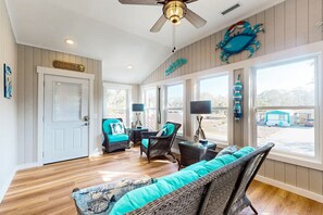 A spacious living room with wicker furniture, large windows, and a ceiling fan. - Dog-friendly coastal home with large yard and close to beach (Supply)