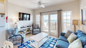 A living room with a ceiling fan, a TV, a sofa, a coffee table, and a view of the ocean through large windows. - Darling oceanfront condo with outdoor pool, hot tub, balcony & views (Hatteras)