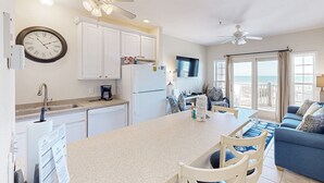 A modern kitchen with white cabinets, a central island, and a dining area with a blue sofa. - Darling oceanfront condo with outdoor pool, hot tub, balcony & views (Hatteras)