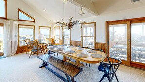 Un vaste espace dédié à la salle à manger, avec une longue table en bois, de nombreuses chaises et un banc. De grandes fenêtres laissent entrer la lumière naturelle et offrent une vue sur l’extérieur.