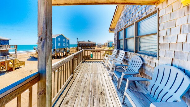 A wooden deck with Adirondack chairs overlooking a beach and a blue building with shutters.
