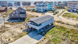 Uma área residencial costeira com casas elevadas em estacas, vista clara da praia e um caminho de areia que leva à água.