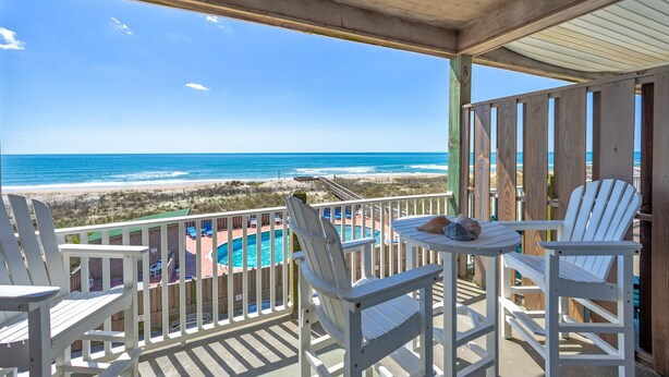 Une terrasse en bois avec des chaises blanches et une table, donnant sur une plage et l’océan.