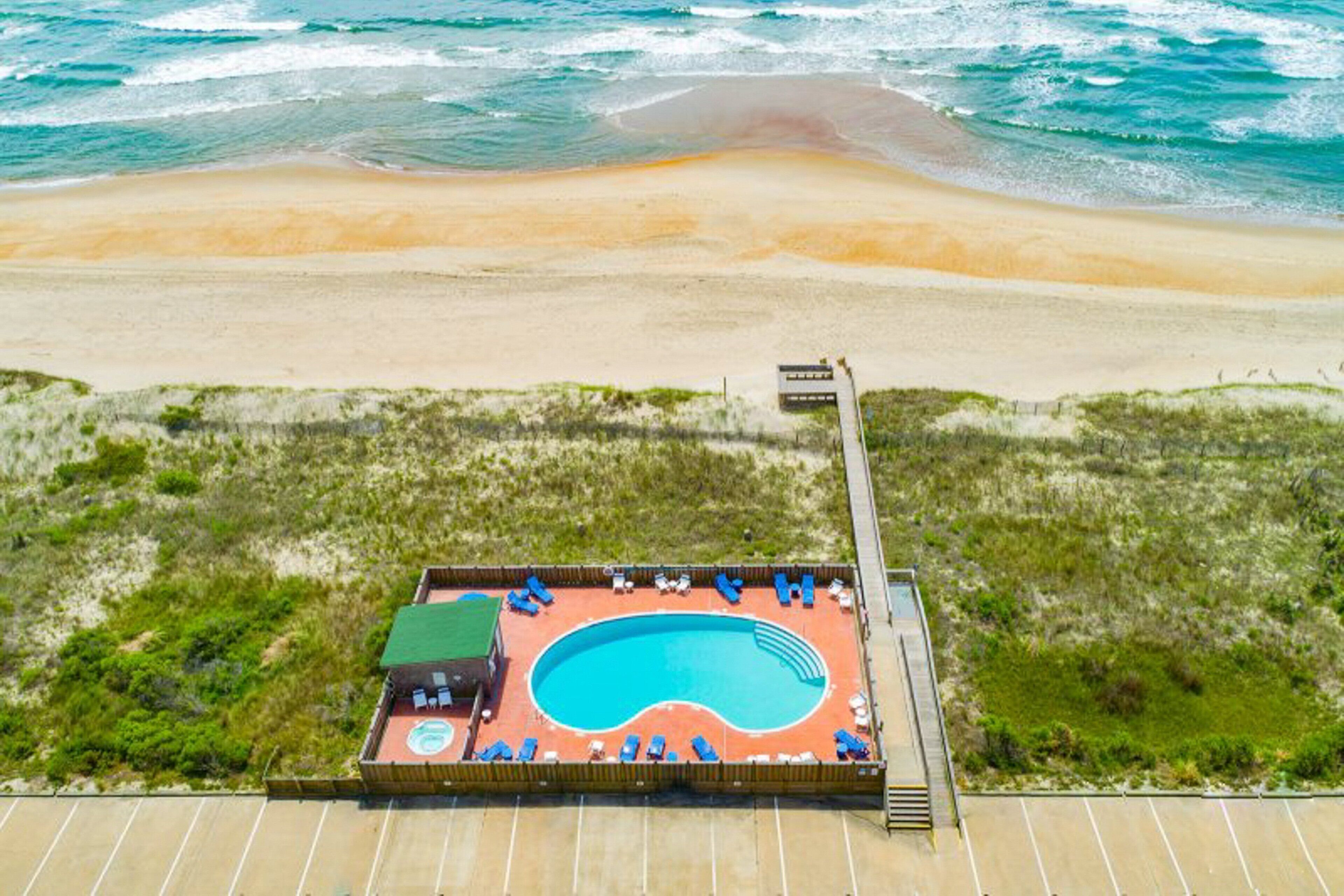 Ovenfra ses en strand med en swimmingpool omgivet af liggestole.