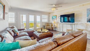 A spacious living room with a large brown leather sectional sofa, a flat-screen TV mounted on a wooden wall, and a ceiling fan.
