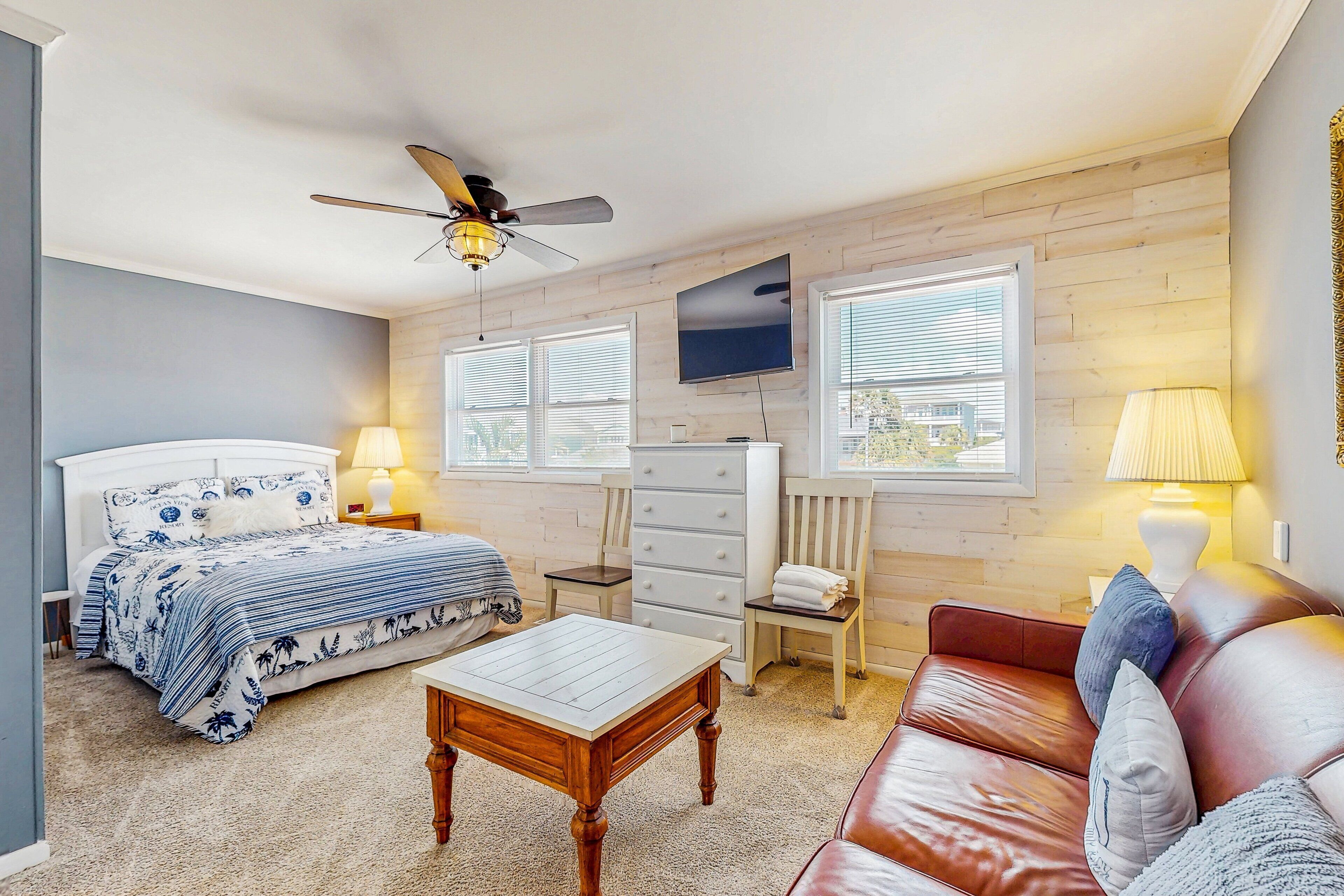 A bedroom with a bed, a wooden table, a leather sofa, a dresser, and a ceiling fan.