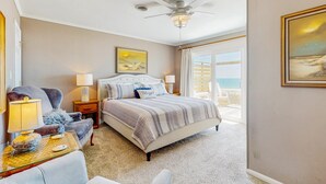 A bedroom with a bed, a chair, a desk, a ceiling fan, and a view of the ocean.