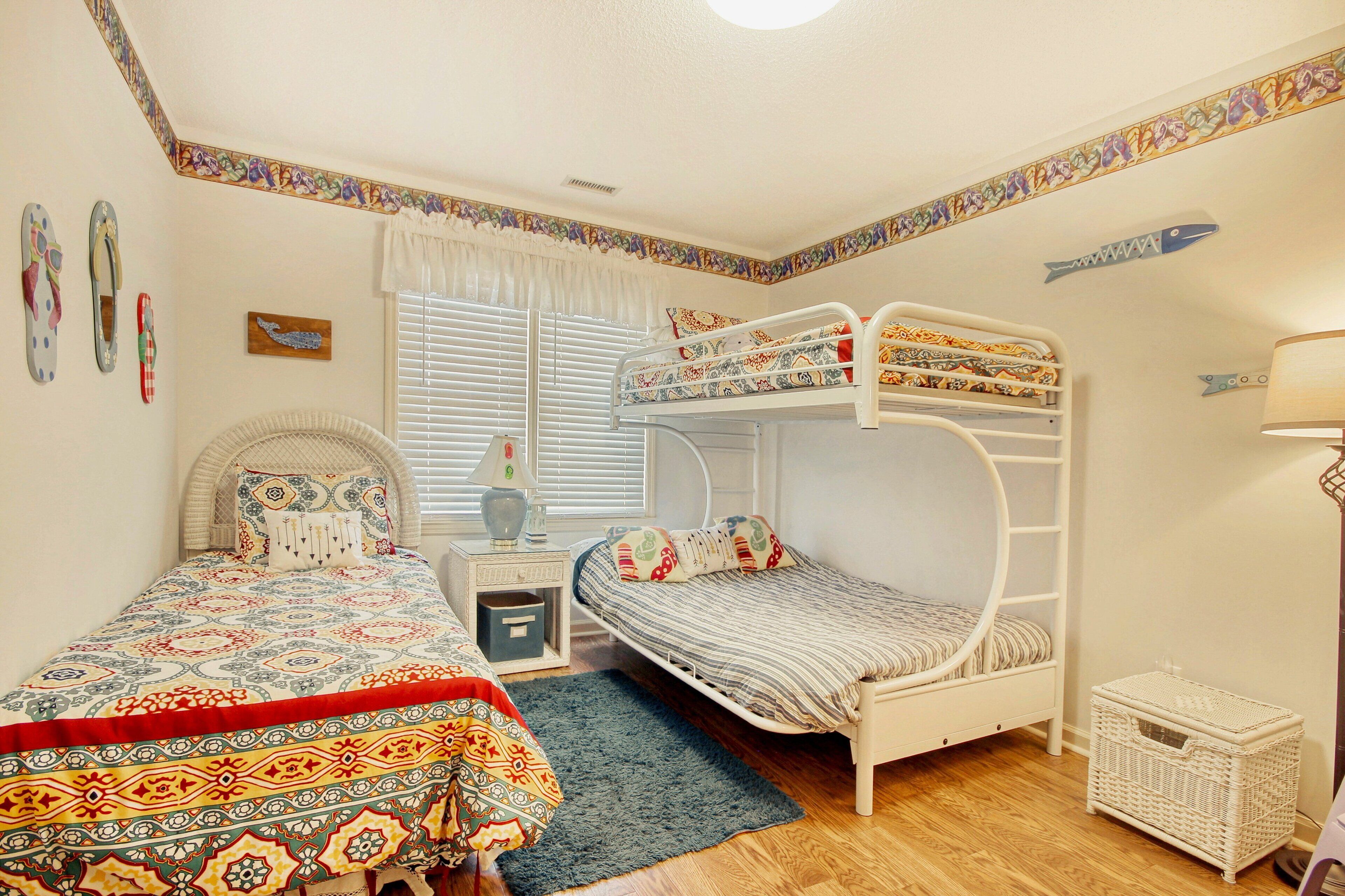 A room with a bunk bed, a single bed, a bedside table, a lamp, and a small wicker basket.