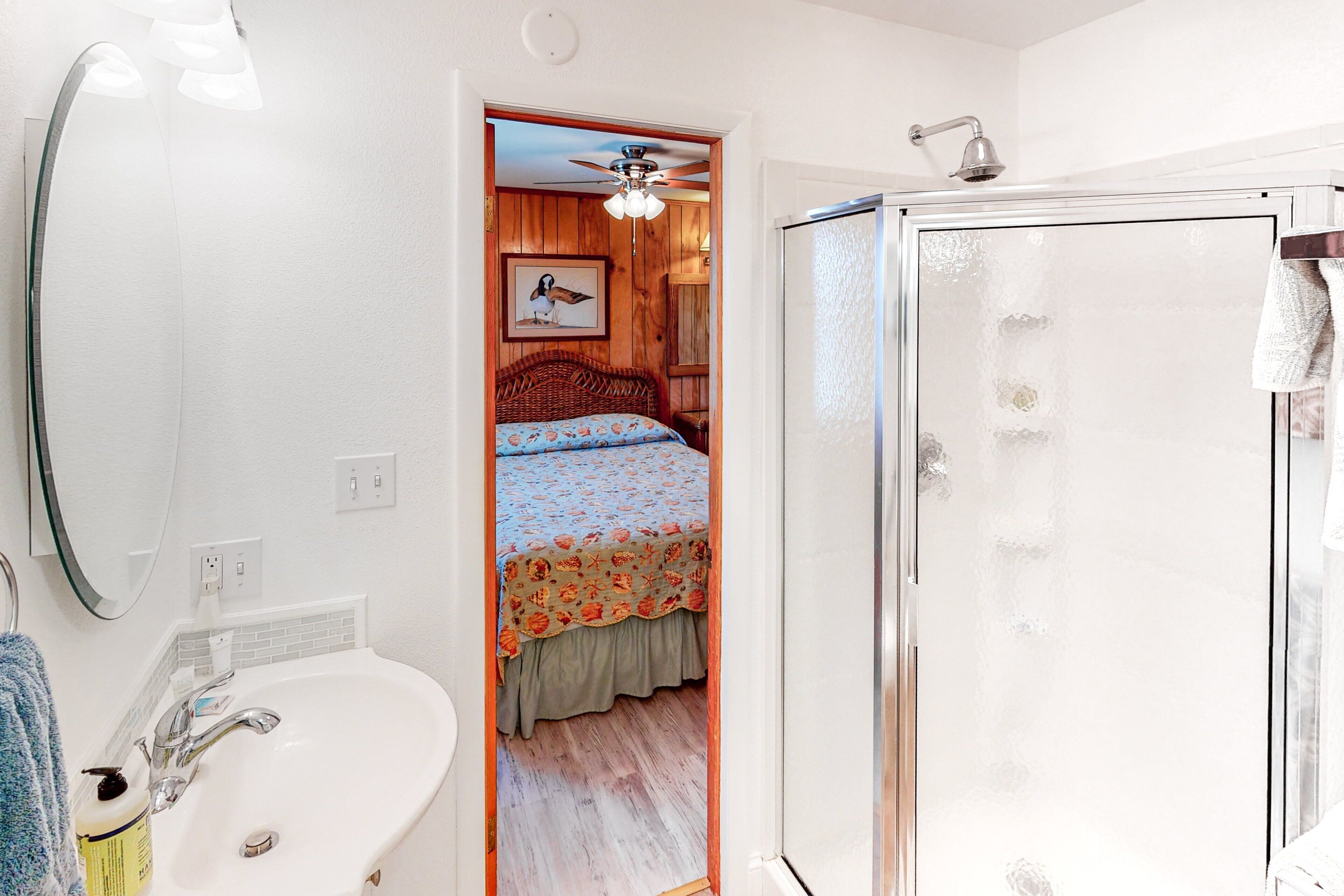 Un baño con un lavabo blanco, cabina de ducha con mampara de vidrio y puerta de madera que da a un dormitorio con cama y ventilador de techo.