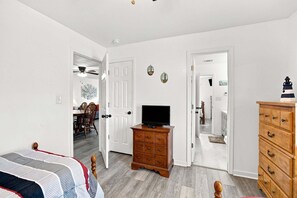 A bedroom with a bed, dresser, TV stand, and a hallway leading to a dining area.