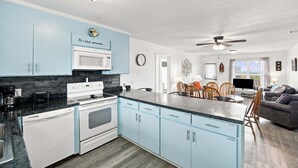 Een moderne keuken met lichtblauwe kastjes, een magnetron, fornuis en vaatwasser. Er is een zithoek met een tafel en stoelen, en een woonkamer met een bank en een televisie.