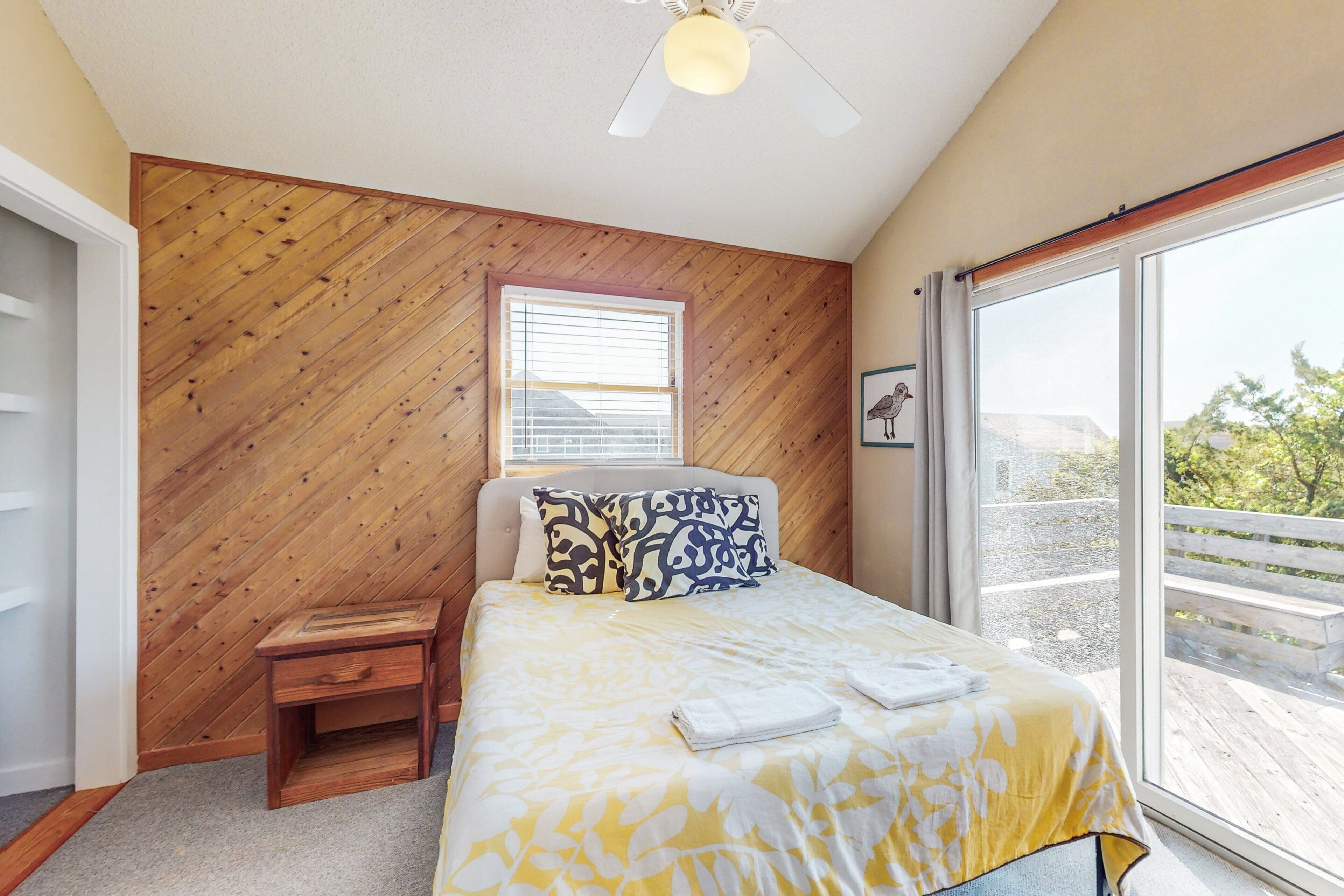 A bedroom with a wooden wall, a bed with a yellow and white bedspread, a nightstand, a ceiling fan, and a large window with a view of a balcony.
