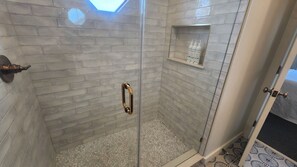 A glass-enclosed shower with a tiled wall, a shelf with toiletries, and a door with a handle. - Ocean-view home with amazing sunset vista, pool access, steps to beach boardwalk (Avon)