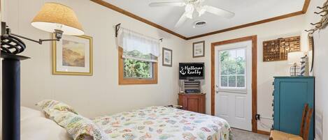 Un dormitorio con cama, ventilador de techo, lámpara, ventana con vistas, puerta y un mueble pequeño.