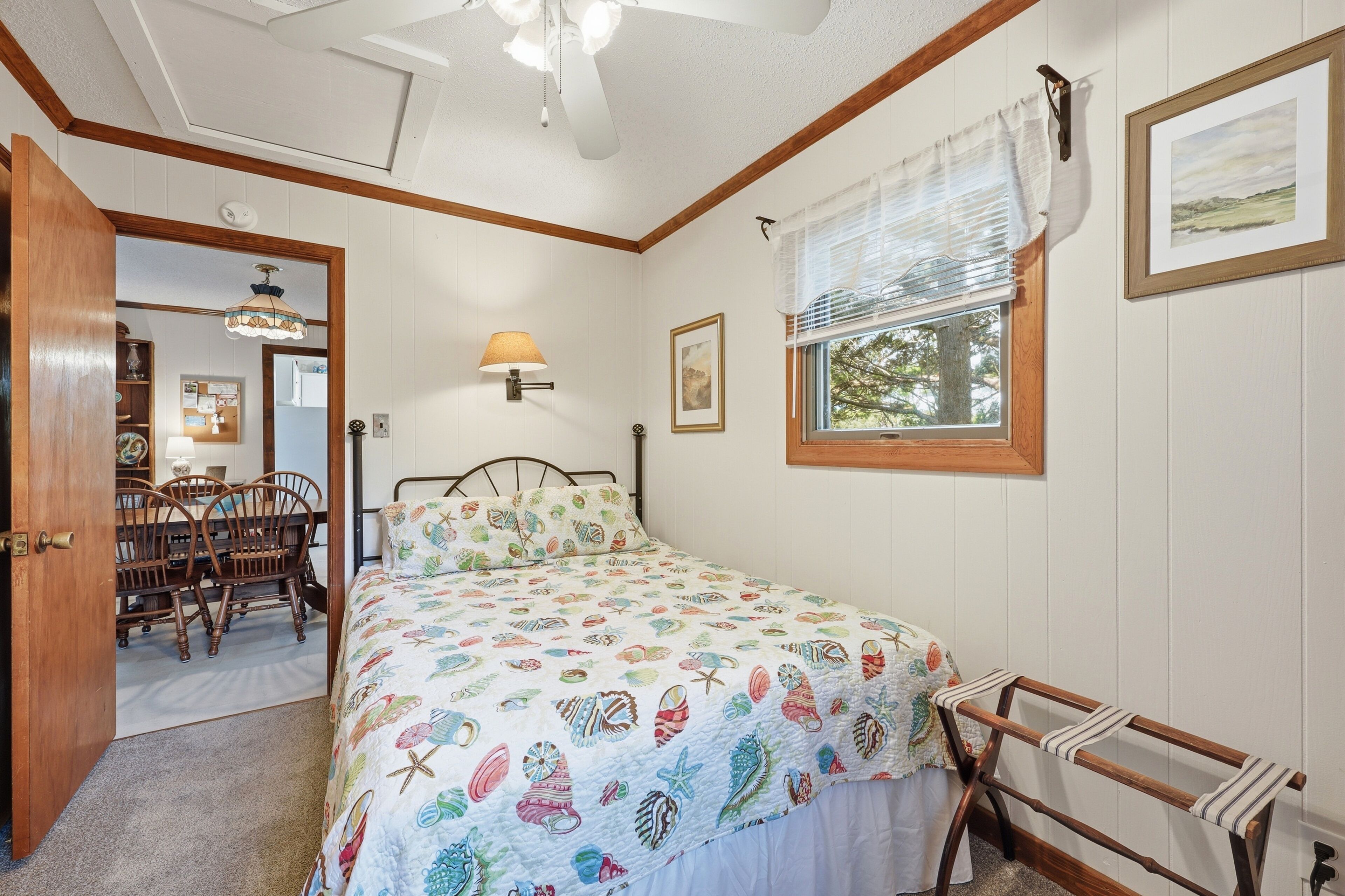 Un dormitorio con cama, ventana con persianas, lámpara de noche, silla de madera y puerta que da a un comedor.