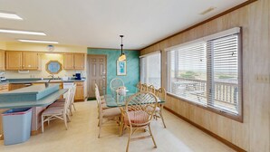 Una cocina con armarios de madera, una zona de comedor con mesa de vidrio y sillas de mimbre, y vistas a través de ventanas amplias con persianas.