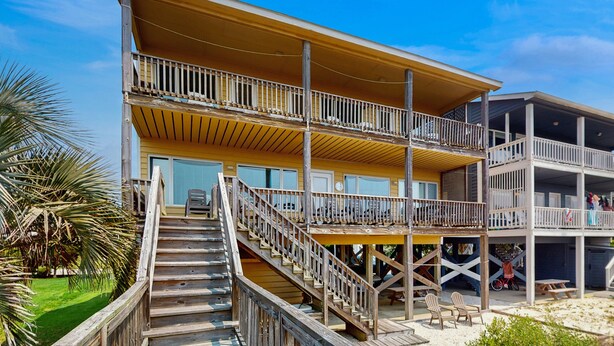 Un edificio frente a la playa con varios pisos, una terraza de madera, asientos al aire libre y una escalera que sube a los niveles superiores.