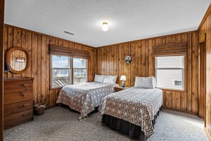 Une chambre avec deux lits, des murs en bois, une commode et une fenêtre avec des stores.