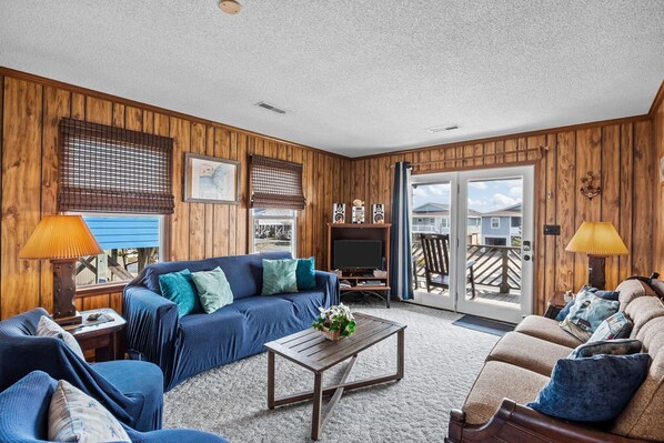 A cozy living room with wooden paneling, a blue sofa, a wooden coffee table, and a television. - Beautiful Home Close to Beach w/Free WiFi, Central AC, Private W/D, Water Views (Holden Beach)