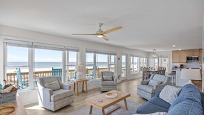 Un salón amplio con un ventanal grande con vistas a la playa, un ventilador de techo y una zona de cocina.