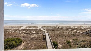 Vista de la playa con un paseo de madera que lleva al agua, dunas de arena y cielo despejado.